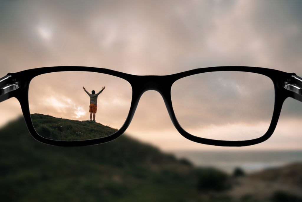 A pair of glasses in front of a landscape scene. Blurred image outside of glasses with clear image looking through glasses of person on hill.